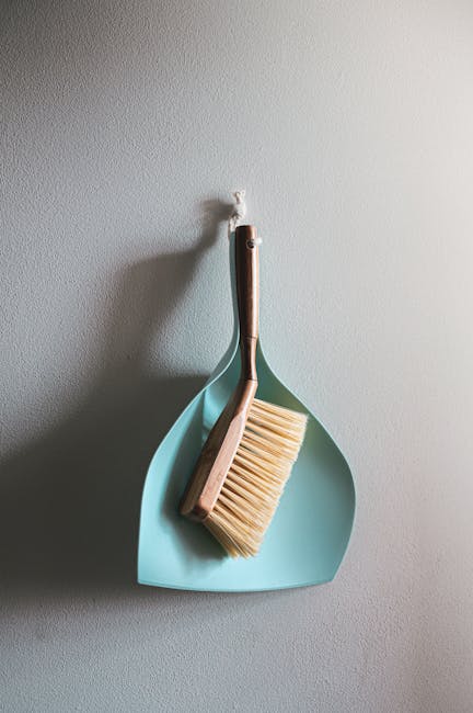 A light blue dustpan hangs on a plain white wall, with a wooden-handled broom and a beige scrubbing brush resting inside it. The broom has natural bristles and is positioned with the handle pointing u