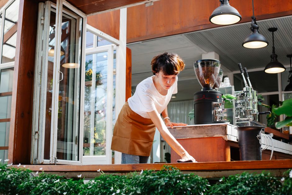A woman with short brown hair wearing a white shirt and a brown apron is leaning over a wooden counter in a bright indoor café or shop, using a cloth to wipe the surface. The countertop holds a commer
