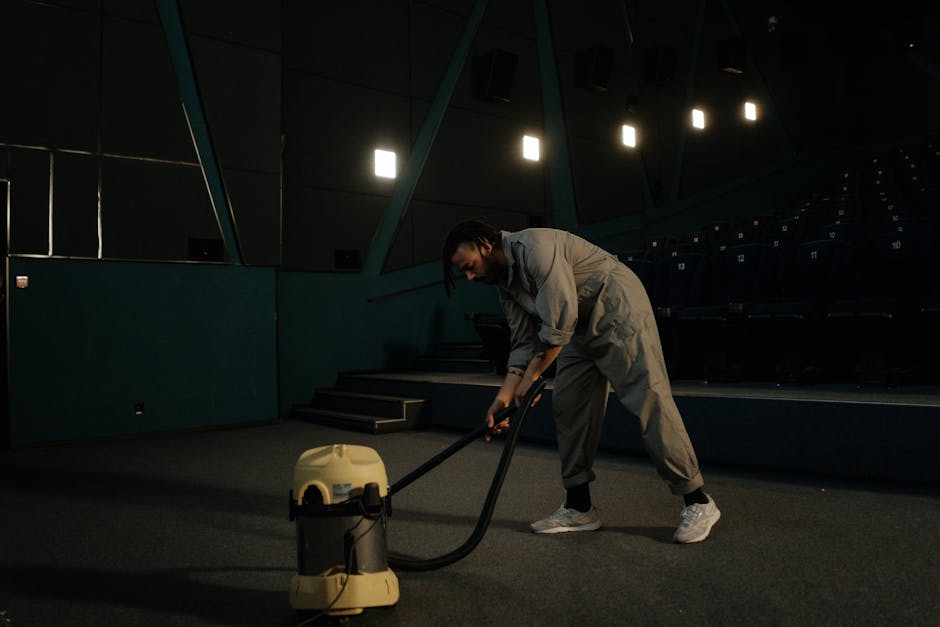 A professional cleaner dressed in a white protective suit, face mask, and gloves, is performing deep cleaning on a plush beige carpet in a well-lit, modern living room. The cleaner is using a steam or